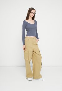 Bluză albastră cu mâneci lungi și pantaloni cargo bej deschis cu buzunare mari pe lateral, asortați cu adidași albi, pe un fundal simplu.