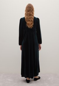 Robe longue en velours vert foncé avec des manches longues et une taille froncée, associée à des chaussures noires. Texture lisse, design à longueur complète.