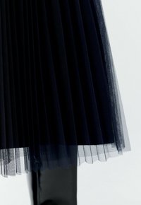 Jupe plissée navy avec un superposé en tulle transparent, matériau lisse et design à plusieurs couches avec des textures contrastées au niveau de l'ourlet.