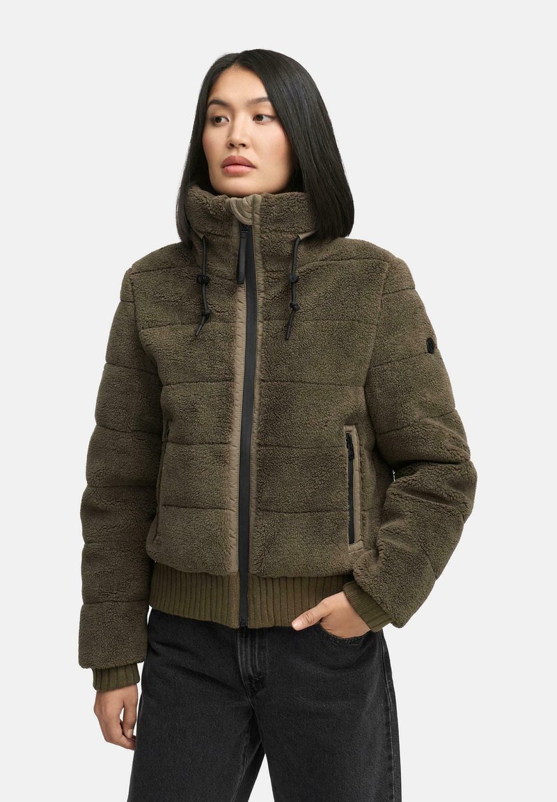 Vrouw in een groene fleecedetail met rits en hoge kraag, met zwarte trekkoorden, staand met één hand in de zak en kijkend naar links.