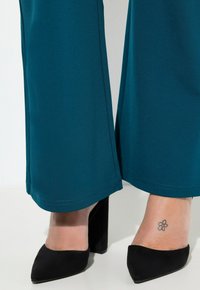 Pantalones de pierna ancha en tono verde azulado con una textura suave, con un dobladillo limpio. Combinados con zapatos de tacón alto negros de punta afilada.