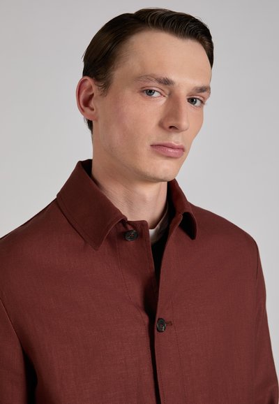 Młody mężczyzna o zaczesanych do tyłu brązowych włosach, ubrany w zapinany na guziki ciemnoczerwony płaszcz, na tle jednolitego jasnoszarego tła.