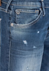 Primer plano de unos vaqueros de mezclilla azul que muestran una textura desgastada, detalles deshilachados, un bolsillo para monedas, presillas para el cinturón y costuras en la zona del muslo.