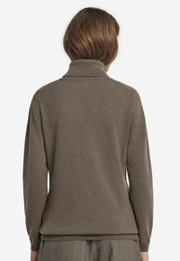 Brun polokrage-sweater som bärs av en person sedd bakifrån, ihop med grå byxor mot en enkel vit bakgrund.