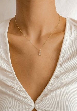 Goldkette mit einem kleinen rechteckigen Anhänger, der klare Steine zeigt. Getragen auf dem Schlüsselbein über einem weißen strukturierten Oberteil.