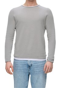Homme portant un pull à manches longues gris clair texturé sur un sous-vêtement blanc et un jean en denim bleu, se tenant avec les bras relaxés.