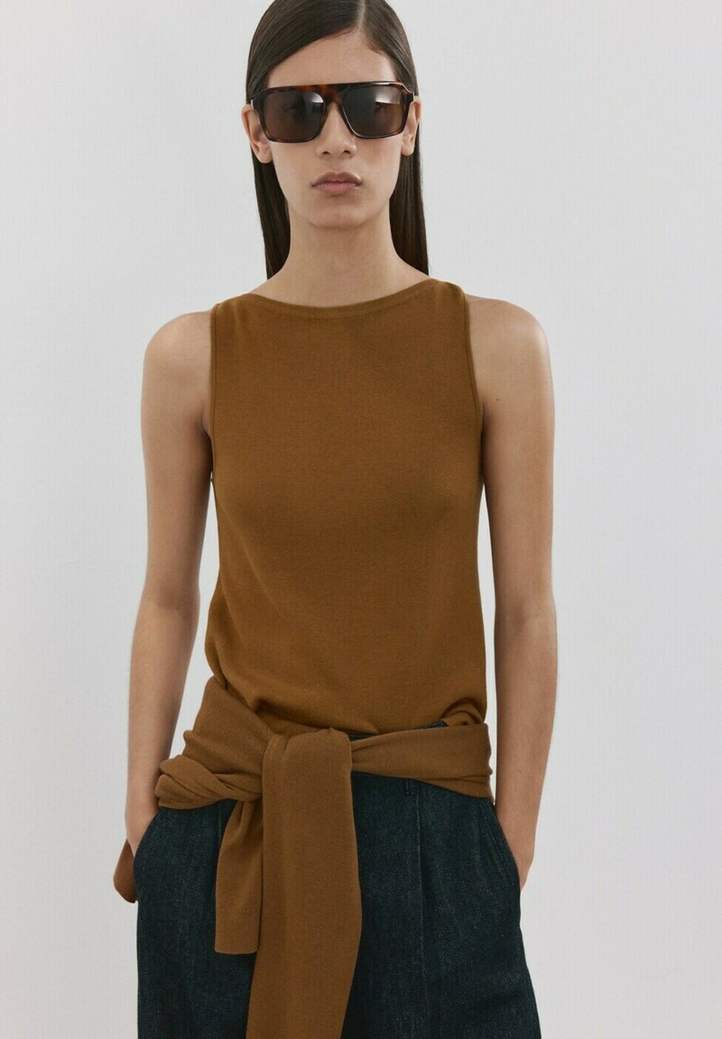 Kobieta ubrana w brązowy top bez rękawów z dopasowanym swetrem związanym w talii, ciemne spodnie i duże okulary przeciwsłoneczne w oprawkach w kolorze żółwia na jednolitym tle.