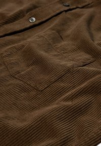 Tessuto in corduroy marrone con strisce verticali, dotato di una tasca a toppa e bottoni in metallo, che crea un aspetto morbido e texturizzato.