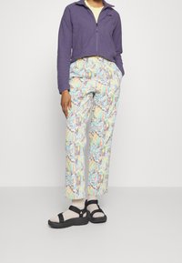 Fioletowa polarowa kurtka na zamek, wielokolorowe spiczaste spodnie w wzory oraz czarne sandały z dwoma paskami, noszone w beżowych skarpetkach.