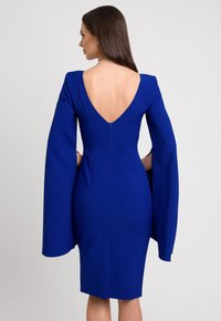 Niebieska sukienka z głębokim dekoltem z tyłu, dopasowanym kroju i długimi, rozszerzanymi rękawami. Materiał wydaje się gładki z delikatnym połyskiem, uwydatniony przez ukryty zamek błyskawiczny.