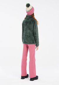 Giacca in pile verde e soffice con colletto alto, abbinata a pantaloni rosa e un cappello crema con pom-pom e dettagli a righe. Calzature nere.