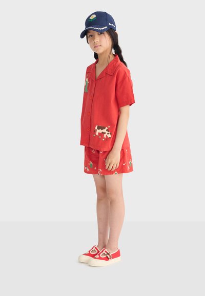 Fille portant une chemise rouge à manches courtes boutonnée et un short avec des imprimés d'animaux de ferme, une casquette bleue marine et des chaussures rouges avec un détail fleur.