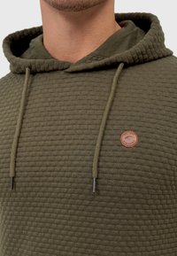 Olijfgroene hoodie van textiel, voorzien van een buidelzak, trekkoorden en een ronde leren logo patch op de borst.