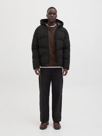 Czarna kurtka puchowa z kapturem, pikowana, zestawiona z brązowym swetrem i luźnymi czarnymi spodniami. Brązowe buty uzupełniają cały zestaw.