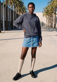 Jeune personne debout à l'extérieur portant un sweat à capuche sombre, une jupe en jean, des chaussettes beige montantes jusqu'aux genoux et des baskets noires avec des palmiers en arrière-plan.