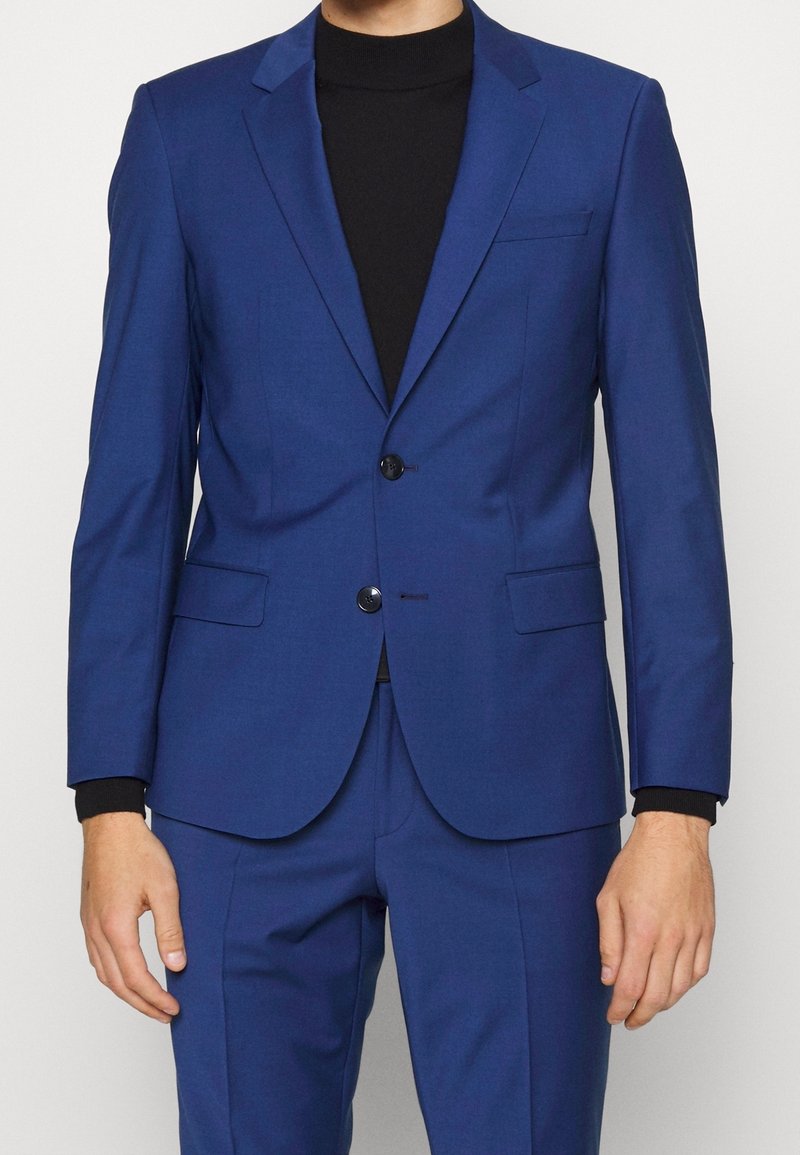 Blauwe outfit met een strak ontwerp, twee knopen, notch revers en twee voorzakken. Gekozen boven een zwart coltrui. Soepele textuur.