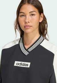 Sweat-shirt Adidas avec un col en V, un corps gris foncé, des épaules blanches et un patch logo texturé. Rayures horizontales sur le col.