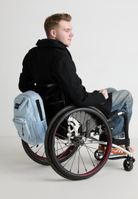 Light blue backpack with mesh accents rests on a black wheelchair. The person wears a black coat and blue jeans, with orange shoes visible.