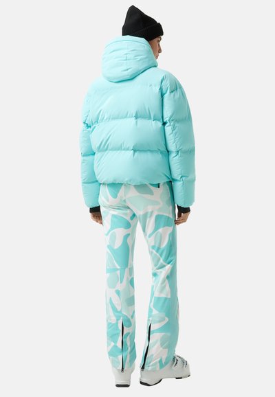 Jasnoniebieska kurtka puchowa z kapturem, w zestawie z turkusowymi spodniami moro z zamkami przy kostkach. Model odwrócony tyłem.