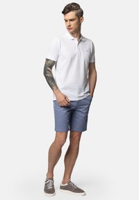 Biała koszulka polo, jasnoniebieskie szorty, szare tenisówki. Model stoi z rękami w kieszeniach, prezentując tatuaż na lewym ramieniu. Prosty design.