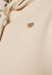 Beige mikina s kapucí a měkkou texturou, s malým vyšívaným zlatým srdcem na levé straně hrudi. Stahovací šňůrka u kapuce.