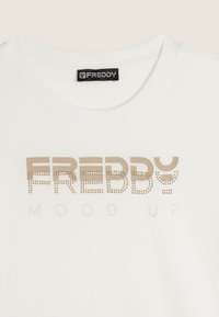 Camiseta de algodón blanca con el logo "FREDDY" en beige, decorada con pequeños remaches. Cuello redondo y mangas cortas. Textura suave.