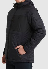Schwarze gesteppte Jacke mit Kapuze, die ein kontrastierendes schwarzes Panel auf der Brust, Reißverschlusstaschen und eine strukturierte Stoffoberfläche aufweist.