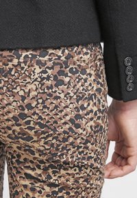 Pantalon à imprimé léopard porté avec une manche de veste noire comportant quatre boutons noirs, et une main reposant près de la hanche.