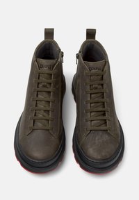Un paio di sneakers alta in pelle verde oliva scuro con suole nere e battistrada rosso, allacciate, viste dall'alto su uno sfondo bianco.