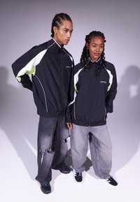 Casacos corta-vento pretos com detalhes em branco e verde néon, combinados com jeans largos e desgastados em tons de cinza. Adequados para uso casual.