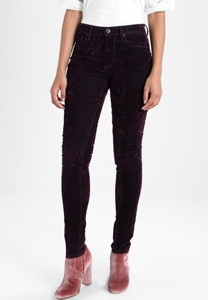 Kobieta ubrana w ciemnofioletowe, aksamitne, obcisłe spodnie z guzikami i zamkiem oraz różowe aksamitne botki na klockowym obcasie, stojąca na białym tle.