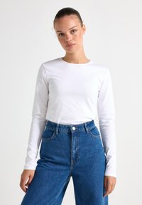 Vit långärmad t-shirt med rund halsringning, matchad med högmidjade blå denimbyxor med vida ben som har framfickor och knappstängning.