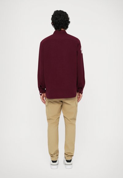 Bordeaux langermet skjorte med knappeliv og en sydd design på venstre erme, kombinert med beige cargo-bukser og hvite joggesko.
