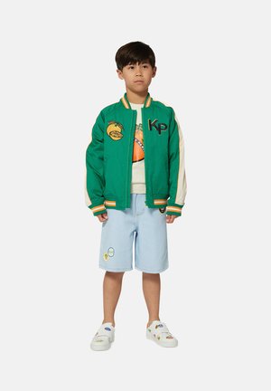 Veste bomber verte avec des accents orange et blanc, ornée de broderies de fruits. Short bleu clair avec des patches et chaussures à enfiler avec des motifs colorés.