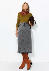 Un haut à manches longues vert olive associé à une jupe en denim grise taille haute. Des bottes en suède marron jusqu'au genou et une écharpe à motifs marron complètent la tenue.