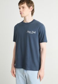 Marineblauwe katoenen t-shirt met korte mouwen, ronde halslijn en "New York" in het wit geborduurd op de linkerkant van de borst. Gladde textuur.