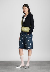 Grønn crossover-veske med preget tekst, brukt over en svart ribbet cardigan, parret med denimshorts med blomsterbroderi og sølvfargede sko.