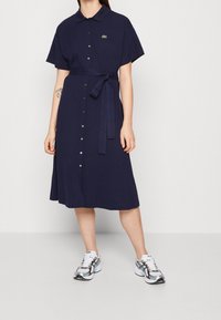 Marinblå klänning med knappar och krage, korta ärmar och ett knytband i midjan. Visar en liten logotyp och en utsvängd nederdel.