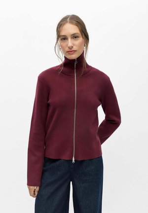 Femme à la peau claire et aux cheveux bruns portant un pull à col roulé bordeaux zippé et un pantalon bleu foncé sur fond blanc uni.
