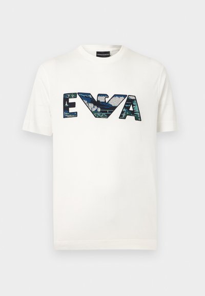 Λευκό κοντομάνικο T-shirt με μεγάλα τυρκουάζ-πράσινα παγιετένια γράμματα "EVA" και ένα στυλιζαρισμένο λογότυπο αετού στο κέντρο.