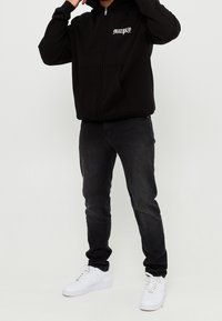 Zwarte zip-up hoodie met voorzakken en logo, gecombineerd met donkere jeans en witte sneakers. Glad materiaal, ontspannen pasvorm.