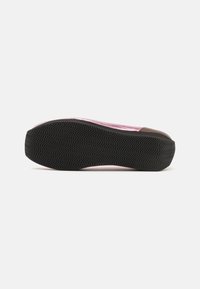 Roza slip-on obuvala z teksturirano zgornjo plastjo in rjavim zaščitnim delom na prstih. Ima črn gumijast podplat z vzorcem cikcak za boljši oprijem.