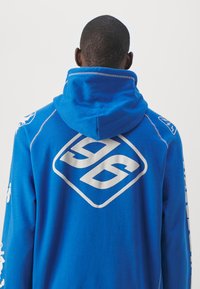 Homme aux cheveux courts portant un sweat à capuche bleu avec un grand logo blanc "96" dans le dos et des détails graphiques blancs sur les manches.