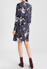 Abito a maniche lunghe di colore blu navy con grande stampa floreale bianca e bordeaux, caratterizzato da un orlo curvo e abbinato a stivaletti con tacco bordeaux.