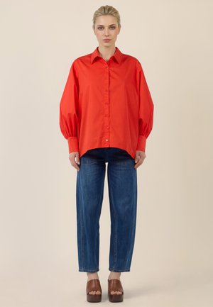 Woman standing wearing a loose red button-up shirt, blue straight-leg jeans, and brown platform open-toe shoes against beige background.