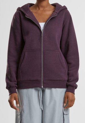 Persona con sudadera con capucha morada con cremallera y bolsillos delanteros sobre una camiseta blanca y pantalones cargo gris claro sobre un fondo liso.