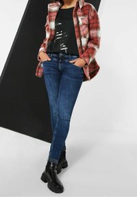 Červená a bílá kostkovaná bunda přes černé tričko s potiskem a tmavé modré úzké džíny. Černé kotníkové boty s robustními podrážkami.