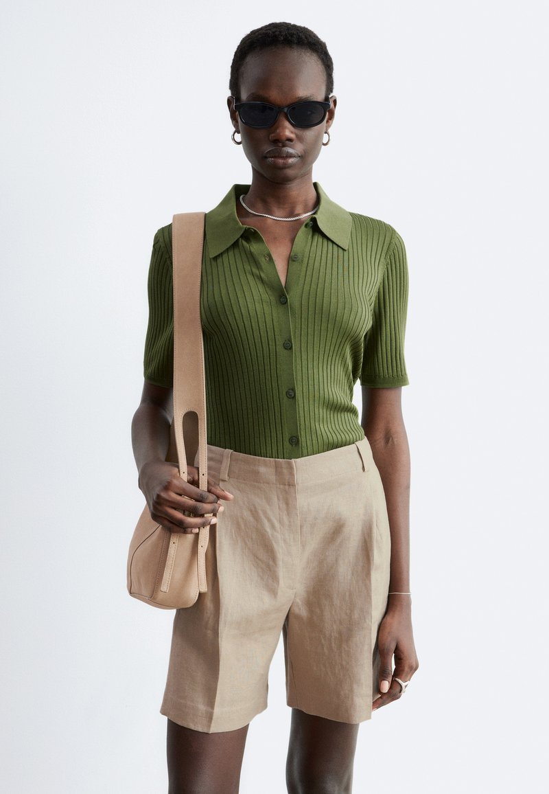Femme portant un haut vert côtelé à manches courtes avec boutons, un short beige taille haute, des lunettes de soleil noires, portant un sac beige à l'épaule.