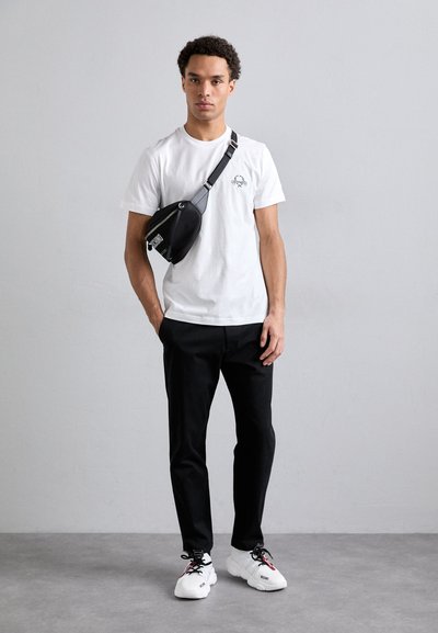 Tricou alb cu un grafic mic, borsetă neagră cu curea, pantaloni negri și adidași albi cu accente de culoare. Fundal simplu.