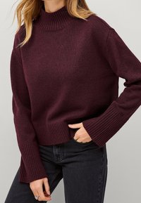 Pull à col roulé en maille bordeaux avec poignets et ourlet côtelés. Texture lisse avec une coupe décontractée ; porté avec un jean noir.